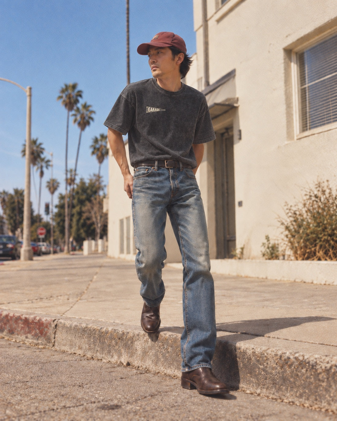 Model wearing the washed-black Garage pennant tee with faded jeans and a burgundy cap on a sunny palm-lined street.